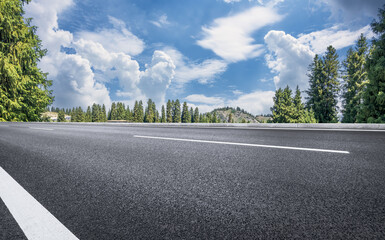 Empty asphalt mountain road highway leads through lush green pine tree forest under blue sky with...