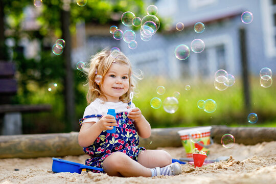 Beautiful little blonde toddler girl having fun with blowing soap bubble blower. Cute adorable baby child playing on playground on sunny summer day. Happy active funny healthy kid