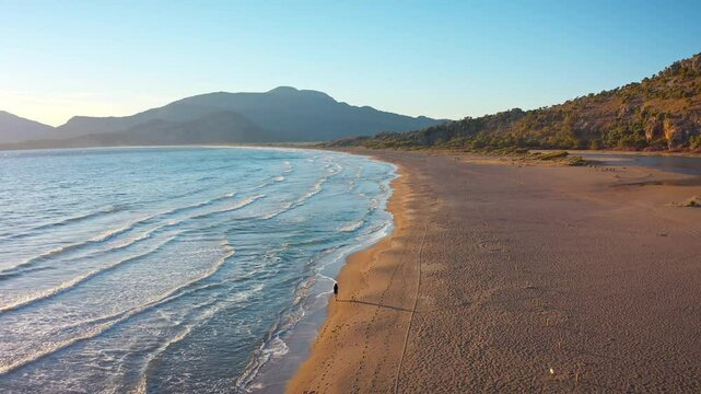 İztuzu Beach drone follows a woman walking along the shore. The sunset is magnificent, the coast and mountains take aesthetics and minimalism to the peak.