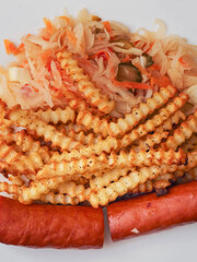A plate of food with a hot dog and French fries on it and sauerkraut salad. The plate is on a wooden table. Tasty dinner.
