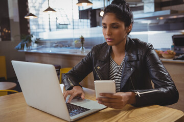 Indian woman typing on laptop and checking smartphone at wooden table in modern café, showing focus