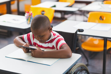 African American boy sitting in wheelchair at school, focusing on tablet and notebook, copy space