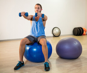 Man sitting on blue stability ball in gym, lifting blue dumbbells with purple exercise ball