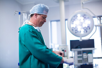 Mid-adult male surgeon pulling on sterile gloves in operating room, with IV bags and surgical light