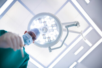 Male surgeon peering down holding scalpel under overhead LED light in operating room, copy space
