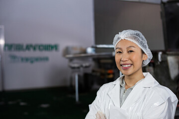 Woman standing, smiling with crossed arms in food processing plant, lab coat, hair net, copy space