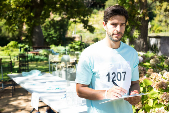 Mid-adult man wearing race bib 1207 standing by folding table in leafy park writing on clipboard