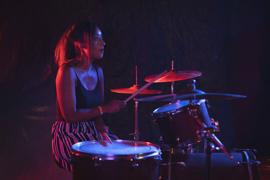 Young adult Asian female drummer performing on stage with drum kit under red and blue lights