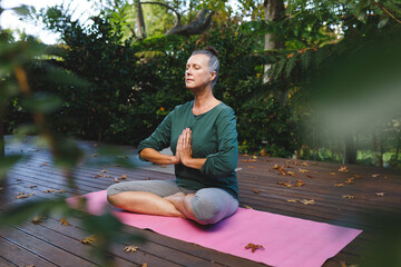 Senior woman sitting cross-legged meditating on pink yoga mat on deck in garden, with autumn leaves