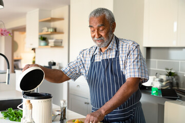 Senior man lifting compost bin lid while chopping bell peppers and herbs at modern kitchen island