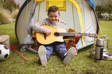 child boy strumming acoustic guitar in backyard, with yellow-gray tent and camping gear