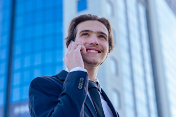 Smiling businessman near a business center calling by cell phone. A happy young businessman talking with a smartphone. Young cheerful businessman near a modern tall glass building with a phone. .