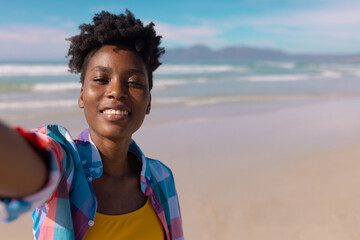 African American woman smiling and taking selfie on sunny sandy beach near ocean waves, copy space