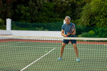 Senior man returning tennis ball on outdoor court, gripping racket near net and protective netting