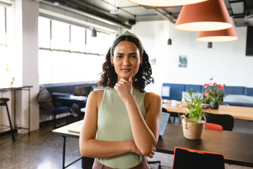 Woman standing thoughtfully in modern coworking office, surrounded by laptop, notebooks and plants