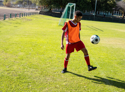 Male athlete juggling soccer ball on grass soccer pitch wearing red bib and cleats, copy space