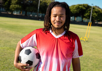 Man wearing red and white jersey holding soccer ball on grass training field with yellow hurdles
