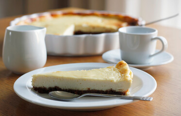 Piece of homemade cheesecake on a white plate on wooden table, a cup of black coffee with saucer, a cup of milk and a baking dish in the background. Coffee nad cake, side view, selective focus.