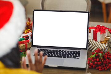 Adult wearing Santa hat waving at silver laptop on wooden table with wrapped gifts at home