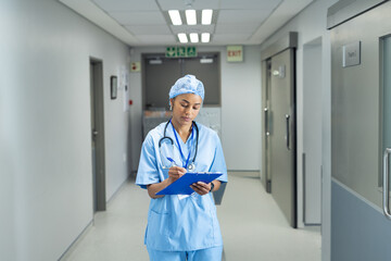 African American woman standing in hospital corridor, writing on blue clipboard with pen