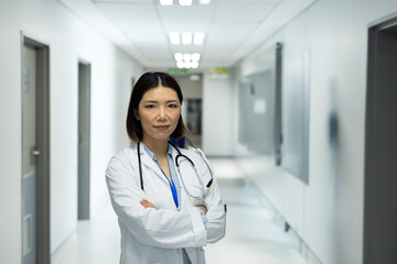 Mid adult Chinese doctor standing arms crossed in hospital corridor, with stethoscope and ID badge