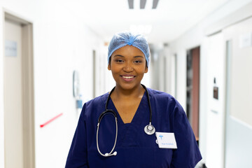 Female nurse smiling and standing in hospital corridor, wearing surgical cap, stethoscope, ID badge