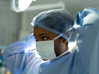 Female surgeon adjusting mask straps and surgical cap in operating room, with bright overhead light
