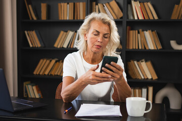 Confident stylish European middle aged senior woman using smartphone at workplace. Stylish older mature 60s lady businesswoman with cell phone in office. Boss leader using internet apps