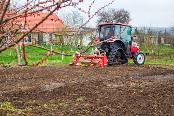 A tractor equipped with a red rotary cultivator actively prepares the soil in a rural field. The...