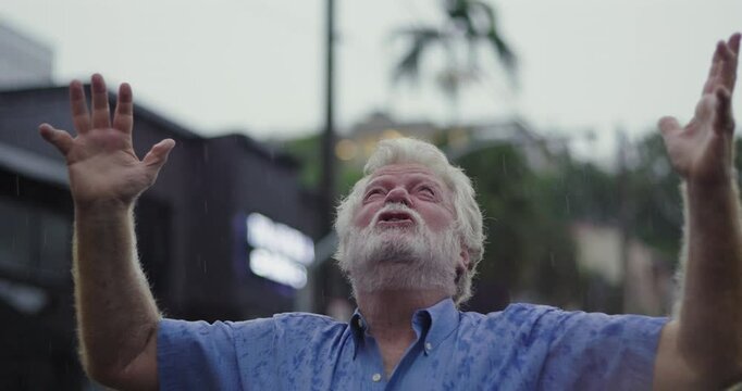 Elderly man with arms stretched wide and face lifted to sky, embracing rain with expression of spiritual release, gratitude, or emotional surrender
