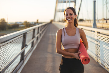 Smiling Woman Holding Yoga Mat on Bridge During Sunrise