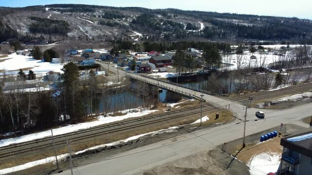 A vehicle crosses a bridge spanning the Matapedia River, which is bordered by a railway, in a small rural village dotted with patches of snow. Sainte-Florence, Quebec, Canada, 2025.