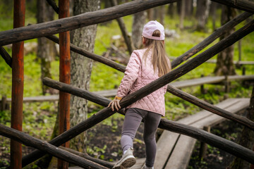 A young girl wearing a pink jacket and cap climbs over rustic wooden beams in a natural park setting, engaged in playful exploration and outdoor adventure.