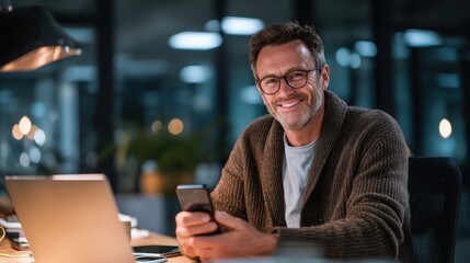 Professional businessman working late, smiling while holding smartphone near laptop on office desk, representing productive evening through digital communication and technology