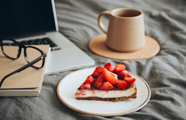 Laptop, coffee cup and piece of strawberry cheesecake on the grey bed sheet.