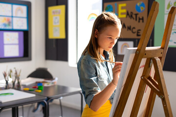 Obraz premium Girl standing by wooden easel in bright art classroom, painting canvas with fine brush, copy space