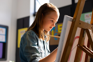 Obraz premium 8-year-old girl painting red circle on canvas at wooden easel in bright art classroom with posters