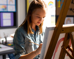 girl painting canvas with red arcs on wooden easel in art studio near window, copy space