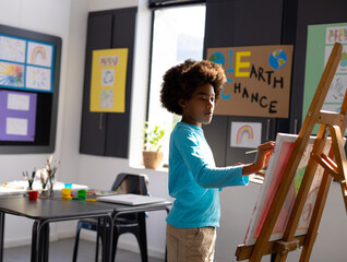 Obraz premium Boy child painting on wooden easel in school art classroom with children's drawings and paintbrush