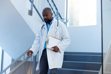 African American male doctor descending hospital stairs, holding clipboard, stethoscope, copy space