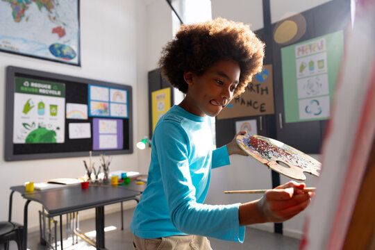 Diverse boy painting on easel in school art studio with paintbrush and palette, copy space