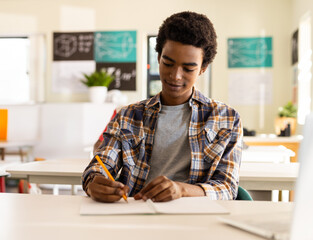 Teenage African American male student sitting in bright classroom, writing notebook with pencil