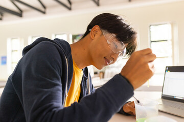 Asian teenage boy pipetting green solution at science lab table with dropper, laptop and goggles