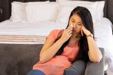 Asian woman sitting on grey sofa in bedroom, wiping face with tissue sadly, copy space