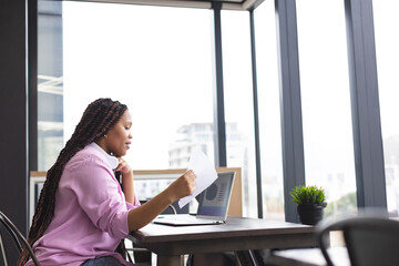 Woman reviewing documents and referencing laptop at table in office with headphones, copy space