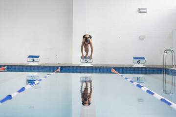 Asian woman swimmer crouching on starting block 5 at indoor pool, with lane ropes, numbered blocks