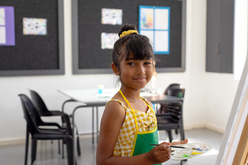 7-year-old girl painting on canvas at easel with palette and brush in art classroom, copy space