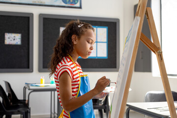 Obraz premium Elementary school girl painting at wooden easel in art classroom, holding paint palette and brush