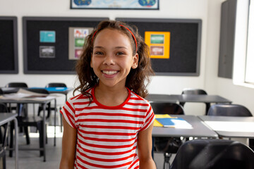 8-year-old girl standing and smiling in school classroom, with desks chairs bulletin board