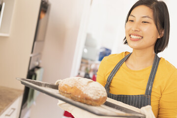 Asian woman holding freshly baked loaf on metal baking tray in home kitchen, copy space